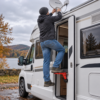 A person uses a folding green car door step to reach the roof of a white motorhome beside an autumn lake.
