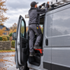A worker stands on an orange type e folding car door step while loading a ladder onto a van roof.