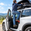 A person uses the green car door step type e green to reach a roof box on a white vehicle through the open door.